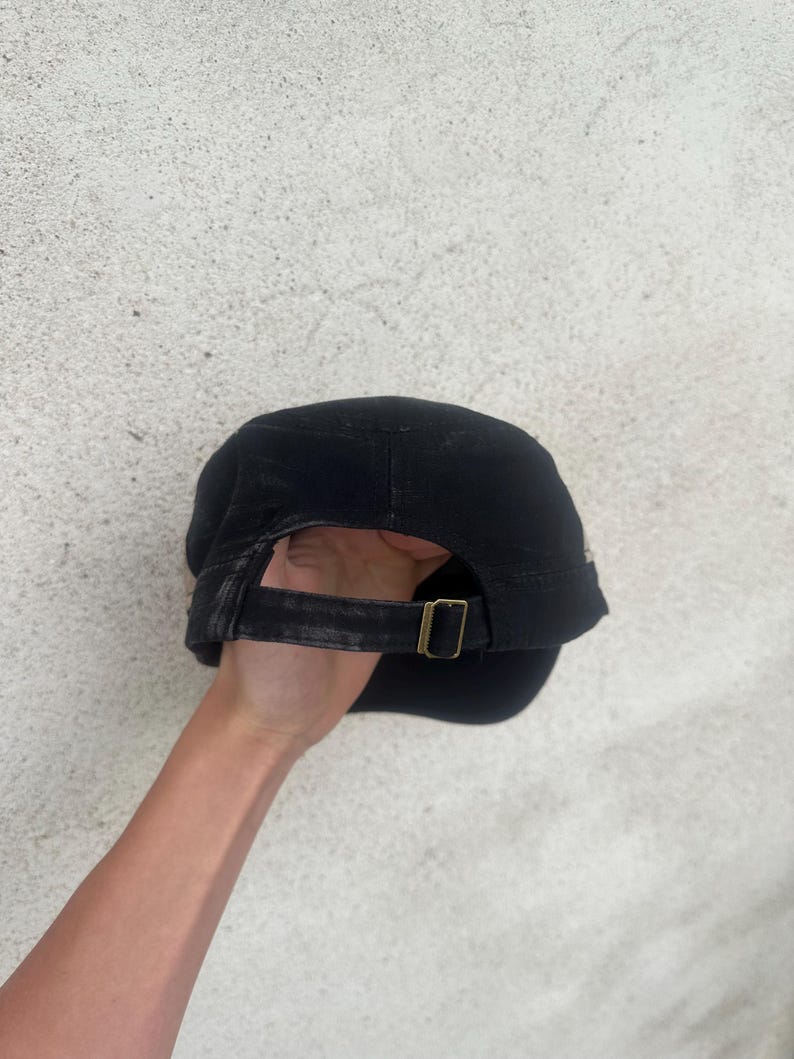 May include: Black cap with a curved brim and adjustable strap. The strap features a brass-colored buckle. The cap is made of a dark fabric and appears to be slightly worn. The cap is a classic style.