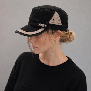 Peut inclure: Casquette noire de style militaire avec un accent beige sur le bord et un triangle beige sur le côté. La casquette a une petite sangle noire avec une étiquette rouge et blanche. La casquette est portée par une personne portant un pull noir.
