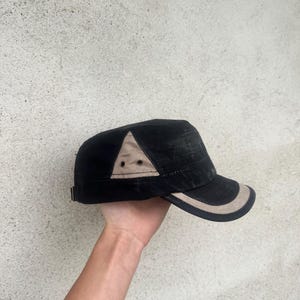 May include: Black and tan military-style cap with a curved brim. The cap features a tan triangle accent on the side and a tan trim along the brim. The cap is held up by a hand.