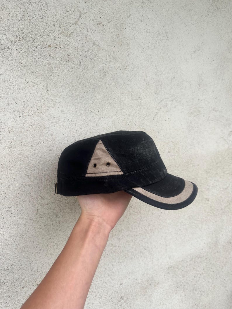 May include: Black and tan military-style cap with a curved brim. The cap features a tan triangle accent on the side and a tan trim along the brim. The cap is held up by a hand.