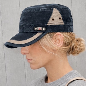 May include: A navy blue military-style cap with a tan accent on the brim and side panel. The cap features a small rectangular patch with the letters "FS" and the word "FASHION". The hat is worn by a person with blonde hair.