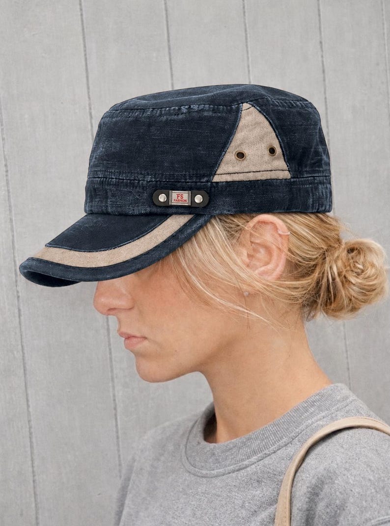 May include: A navy blue military-style cap with a tan accent on the brim and side panel. The cap features a small rectangular patch with the letters "FS" and the word "FASHION". The hat is worn by a person with blonde hair.