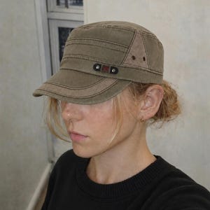 May include: Olive green military-style cap with a short brim. The cap features a rectangular patch with the letters "G&D" and two small rivets. The cap is made of a durable fabric and has a structured design.