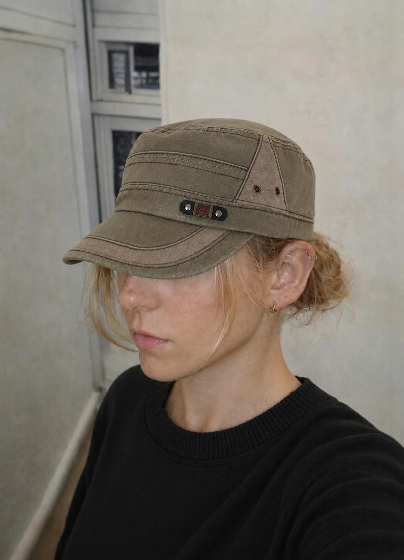 May include: Olive green military-style cap with a short brim. The cap features a rectangular patch with the letters "G&D" and two small rivets. The cap is made of a durable fabric and has a structured design.