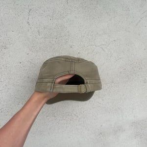 May include: A khaki-colored military-style cap with a curved brim and adjustable strap. The cap has a structured design with visible stitching and a metal buckle. The back of the cap is visible, showing the adjustable strap.