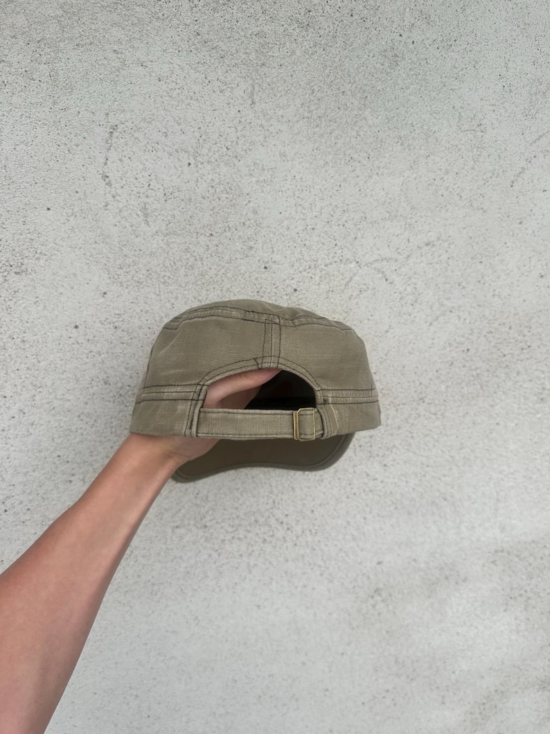 May include: A khaki-colored military-style cap with a curved brim and adjustable strap. The cap has a structured design with visible stitching and a metal buckle. The back of the cap is visible, showing the adjustable strap.
