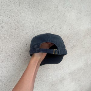 May include: A navy blue military-style cap with a curved brim and adjustable strap. The cap features a brass-colored buckle and a structured crown. The fabric appears to be a durable cotton or similar material, suitable for outdoor wear.
