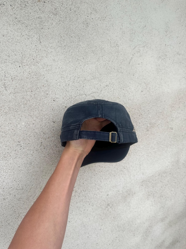 May include: A navy blue military-style cap with a curved brim and adjustable strap. The cap features a brass-colored buckle and a structured crown. The fabric appears to be a durable cotton or similar material, suitable for outdoor wear.