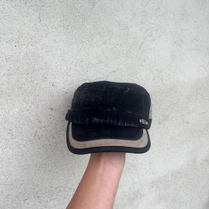 May include: Black military-style cap with a tan brim and accents. The cap has a flat top and a short, curved brim. The cap is held up against a textured, light gray background.