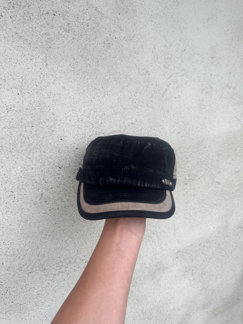 May include: Black military-style cap with a tan brim and accents. The cap has a flat top and a short, curved brim. The cap is held up against a textured, light gray background.