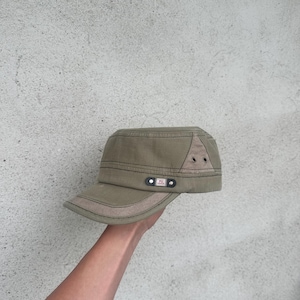 May include: An olive green military-style cap with a short brim. The cap features a tan accent on the brim and a triangular patch on the side with two small eyelets. A small black tag with white text is on the side.