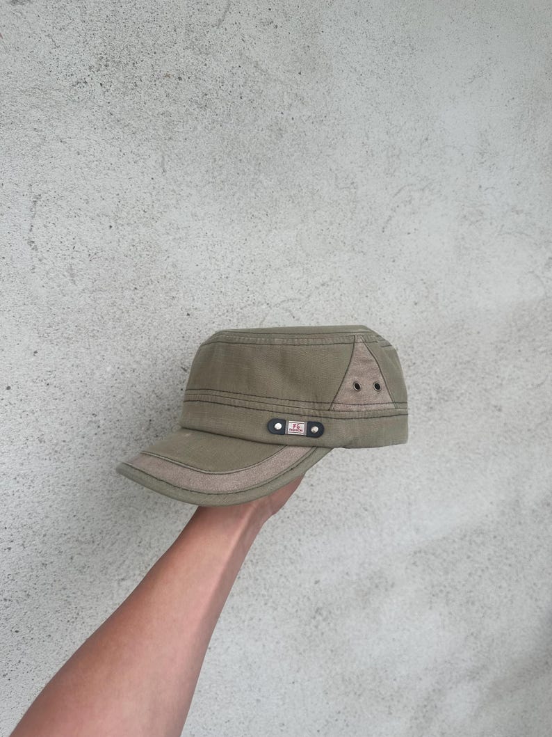 May include: An olive green military-style cap with a short brim. The cap features a tan accent on the brim and a triangular patch on the side with two small eyelets. A small black tag with white text is on the side.