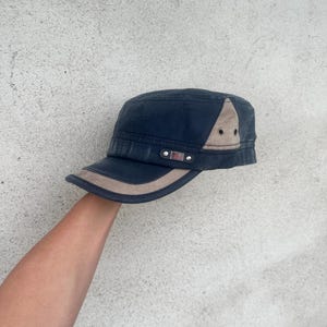 May include: A navy blue military-style cap with a tan brim and accents. The cap features a small American flag patch and a tan triangle on the side. The cap is held up by a person's arm.