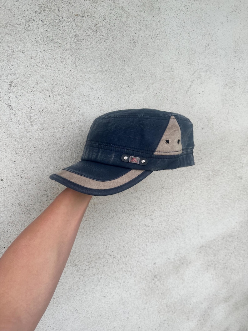 May include: A navy blue military-style cap with a tan brim and accents. The cap features a small American flag patch and a tan triangle on the side. The cap is held up by a person's arm.