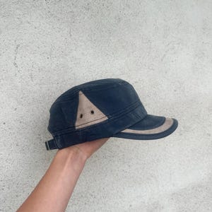 May include: A navy blue cap with a tan accent on the side and brim. The cap has a structured design with a short brim and a metal clasp on the back for adjustment. The cap is held by a person's hand.