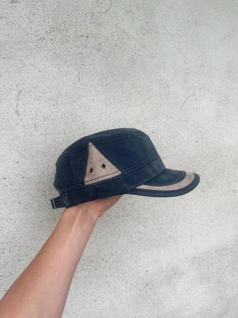 May include: A navy blue cap with a tan accent on the side and brim. The cap has a structured design with a short brim and a metal clasp on the back for adjustment. The cap is held by a person's hand.