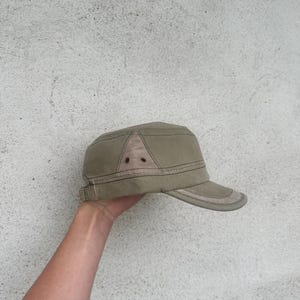 May include: A khaki-colored military-style cap with a short brim. The cap features a structured design with a contrasting fabric panel on the crown and a subtle, adjustable strap. The hat is a neutral accessory.
