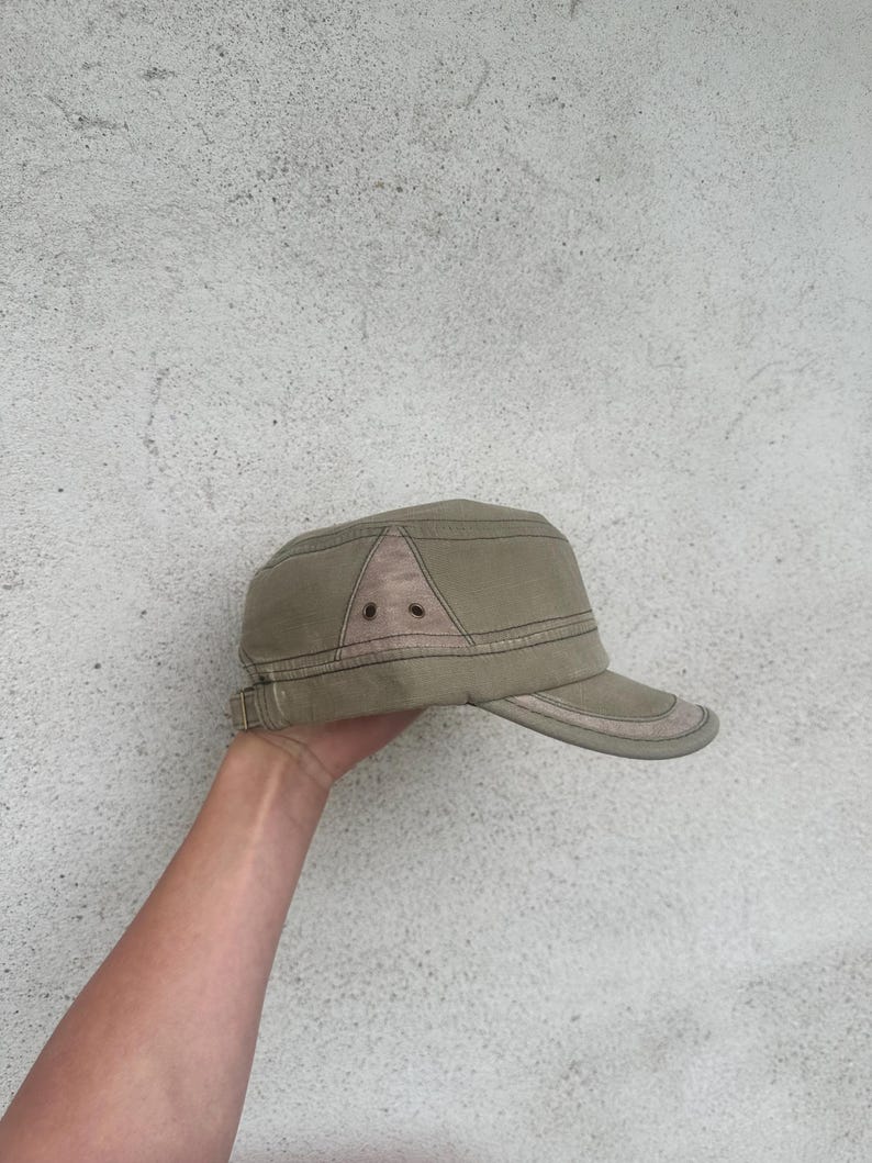 May include: A khaki-colored military-style cap with a short brim. The cap features a structured design with a contrasting fabric panel on the crown and a subtle, adjustable strap. The hat is a neutral accessory.