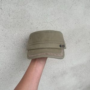 May include: A khaki-colored military-style cap with a flat top and a short, stiff brim. The cap has a structured design with visible stitching and a small, decorative detail on the side. The cap is held up by a person's hand.