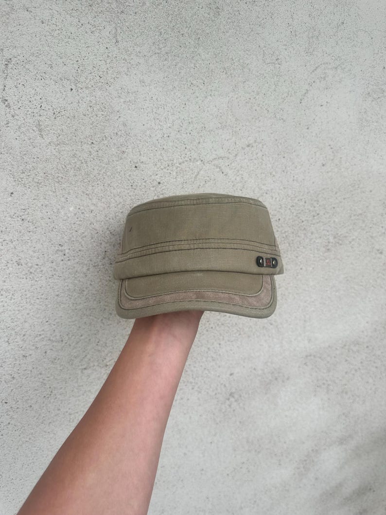 May include: A khaki-colored military-style cap with a flat top and a short, stiff brim. The cap has a structured design with visible stitching and a small, decorative detail on the side. The cap is held up by a person's hand.