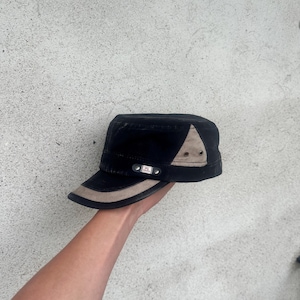 May include: Black military-style cap with a flat brim and a tan accent on the side. The cap has a small brim and is held by a person's hand. The cap has a small metal tag on the front.