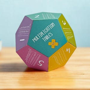 Könnte beinhalten: Ein farbenfrohes Dodekaeder mit Einmaleins-Tabellen auf seinen Flächen. Die geometrische Form ist türkis, lila, gelb und rosa. Das Wort "DIY" befindet sich über dem Dodekaeder, mit einer Schere und einer Klebstoffflasche im Hintergrund.