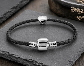 May include: A black braided leather bracelet featuring a polished silver-tone barrel clasp and a single smooth silver-tone heart-shaped charm. The bracelet is circular in shape and displays a high-shine metallic finish on the silver components.