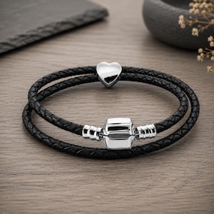 May include: A double-wrap bracelet featuring a black braided cord. The bracelet is adorned with a polished silver-tone heart charm and a central rectangular silver-tone clasp component with ribbed detailing on both sides of the main piece.