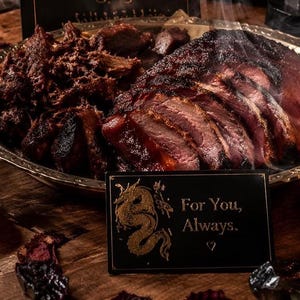 May include: A dark, moody still life featuring a platter of cooked meat, a small lit candle, and a sign that reads "For You, Always." The scene is set on a wooden surface with other decorative elements.