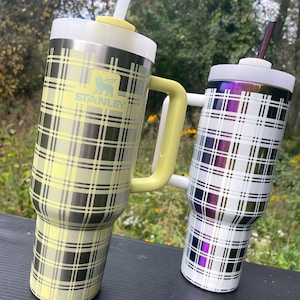 Könnte beinhalten: Zwei Edelstahl-Reisebecher mit karierten Mustern. Der Becher links ist gelb-schwarz kariert mit gelbem Griff und dem Stanley-Logo. Der Becher rechts ist weiß-schwarz kariert mit einem schillernden Finish.