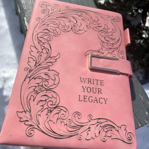 May include: Pink journal with a decorative black floral design on the cover. The journal has a gold-colored clasp and the words "WRITE YOUR LEGACY" printed on the front. A leather strap is visible.