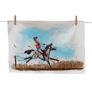 Può includere: Un asciugamano da cucina bianco con un'illustrazione di cartone animato di una persona che cavalca un cavallo attraverso un campo di grano. Il cavallo è grigio e bianco con zoccoli marroni. La persona indossa un gilet rosa e un casco blu. Lo sfondo è un cielo blu con nuvole bianche.