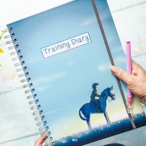 Puede incluir: Una agenda de entrenamiento de espiral azul con una ilustración de dibujos animados de una persona montando a caballo. La portada tiene el texto "Training Diary" en un cuadro blanco.