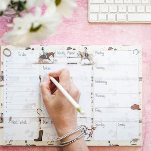 May include: A white weekly planner with a western theme. The planner has a to-do list, notes section, and daily columns for Monday through Sunday. The planner is decorated with cowboys and horses.