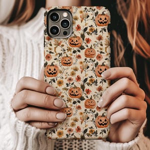 May include: A smartphone case featuring a repeating pattern of orange jack-o'-lanterns with carved faces, interspersed with small yellow and orange flowers, green leaves, and thin brown vines against a light cream-colored background.