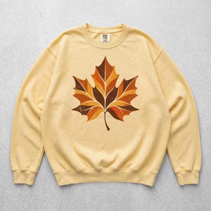 May include: A pale yellow crewneck sweatshirt featuring a large, centered graphic of a maple leaf. The leaf design is composed of segmented, geometric shapes in shades of dark brown, burnt orange, and golden yellow with a distressed texture.