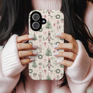 May include: A phone case with a festive Christmas pattern. The design features illustrations of nutcrackers, Christmas trees, wreaths, and wrapped gifts in shades of green, pink, and beige. The case is held by a person wearing a pink sweater.