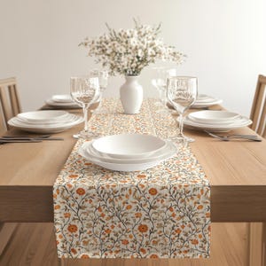 May include: A dining table set for a meal, featuring a floral patterned table runner with orange and brown flowers. White plates, glasses, and a vase with white flowers complete the setting. The table is made of light-colored wood.