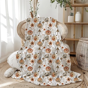 Op de afbeelding: Een witte Halloween-deken met een herhalend patroon van pompoenen, geesten, zonnebloemen en paddenstoelen. De pompoenen hebben uitgesneden gezichten in oranje en bruin. De deken ligt over een rieten stoel.