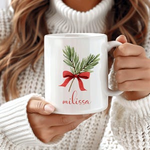May include: White ceramic mug featuring a watercolor design of green pine branches and a red bow. The name "milissa" is written in red script below the design. The mug has a white handle.