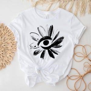 May include: White t-shirt featuring a black and white floral design with an eye in the center. The design is made with brushstrokes, giving it a unique, artistic look. The shirt is tied at the waist.
