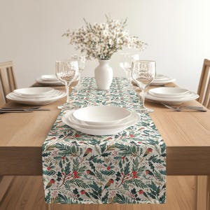 May include: A dining table set for a meal, featuring a patterned table runner with a botanical design of birds, holly, and berries. White plates, glasses, and a vase of flowers complete the setting. The table is made of wood.