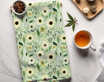 May include: A folded green and white floral patterned kitchen towel with a sunflower design. The towel is decorated with white sunflowers, green leaves, and small yellow flowers. The towel is displayed with a bowl of star anise and a cup of tea.