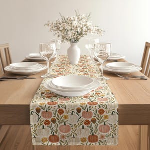 May include: A rectangular table runner featuring a repeating pattern of pumpkins in shades of orange, white, and pink, interspersed with orange and yellow flowers and green foliage, laid flat on a wooden table with white plates and clear glasses.