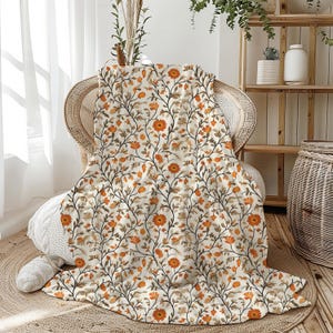 May include: A cream-colored throw blanket with a repeating floral pattern in shades of orange, brown, and gray. The blanket is draped over a wicker chair, creating a cozy and inviting aesthetic. The floral design features stylized flowers and leaves.