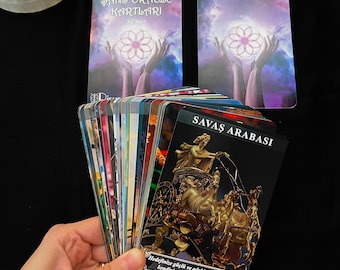 May include: A deck of oracle cards with the title "SANS ORACLE KARTLARI" and "SAVAŞ ARABASI" on the cards. The cards feature illustrations of hands holding a glowing design and a golden chariot. A small beige pouch is also visible.
