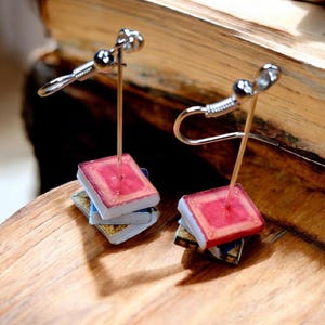 May include: A pair of silver book-shaped earrings with red covers. The earrings are on a stand and are ready to be worn.