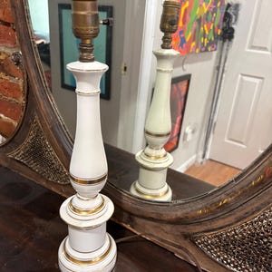 Può includere: Un paio di lampade da tavolo in ceramica bianca con finiture dorate. Le lampade hanno un design classico con un portalampada in alto e una base rotonda. Le lampade sono esposte davanti a uno specchio.