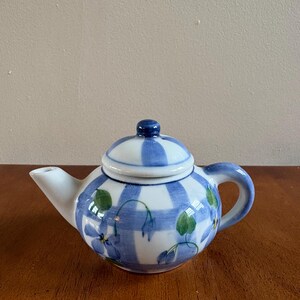 May include: A ceramic teapot featuring a white base with a blue painted plaid pattern. The teapot is decorated with hand-painted blue flowers and green leaves. It includes a matching lid with a small blue knob handle, a curved spout, and a handle.