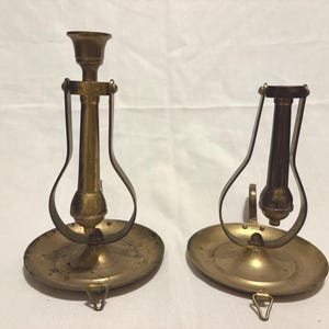 Op de afbeelding: Twee antieke messing kandelaars. Elk heeft een ronde basis, een centrale kolom en een gebogen handvat. De kandelaars zijn gemaakt van messing en hebben een vintage uitstraling. Ze zijn ontworpen voor één enkele kaars.